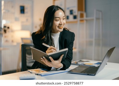 Focused businesswoman smiles and writes in her notebook during a late-night video call, multitasking in a dimly lit office - Powered by Shutterstock