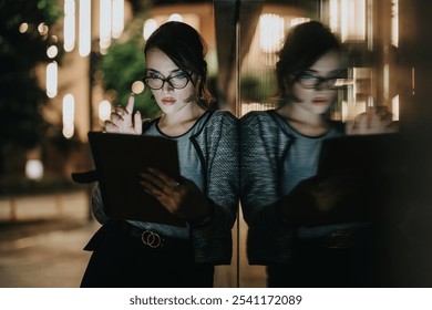 Focused businesswoman using a tablet at night, reflected on a glass wall. Concept of late-night work, technology, and business. - Powered by Shutterstock