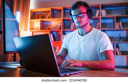 Focused by work. Man in glasses and white shirt is sitting by the laptop in dark room with neon lighting. - Powered by Shutterstock