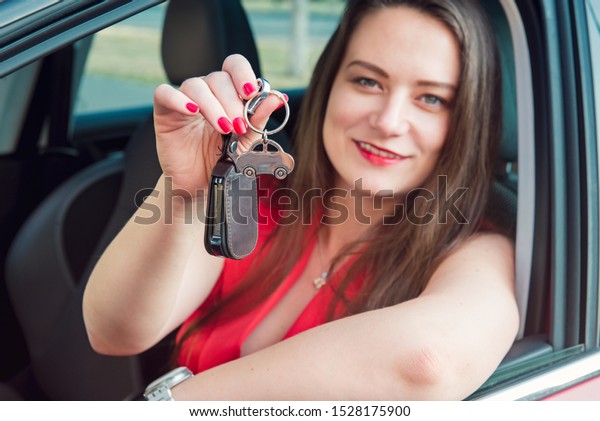 Focused car shaped keychain with keys on the\
background of happy successful woman sitting in her new car.\
Outdoors view. Selective focus, copy\
space