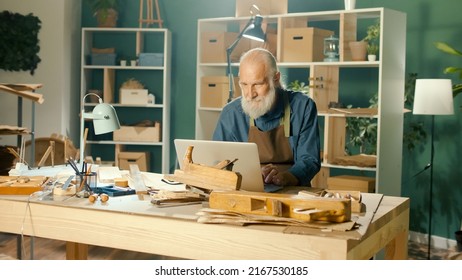 A Focused Carpenter Develops Layouts from Wood, Models 3D Sketches Using a Laptop in a Home Workshop. Garage Hobby, Profession, Joinery and New Computer Technology. - Powered by Shutterstock