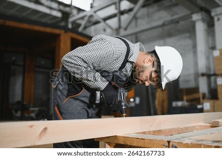 Focused carpenter using cordless drill assembling wooden frame for modular house construction in a bright workshop