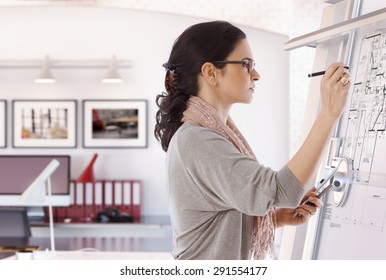 Focused Casual Caucasian Female Architect Working At Drawing Board With Pen In Hand. Wearing Glasses, At Office. Floor Plan, Busy, Concentration, Unsmiling.