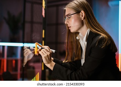 Focused Caucasian Business Woman Writing Task On Sticky Note On Glass Wall, While Developing Project Plan And Strategy In Creative Corporate Office. Female Scrum Master Working In Office