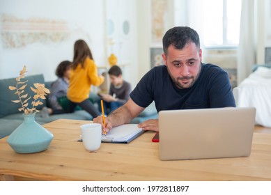 Focused Caucasian Dad Working At Home. Bearded Man Sitting At Table, Looking At Laptop Screen And Writing Information On Paper. Fatherhood, Work At Home Concept