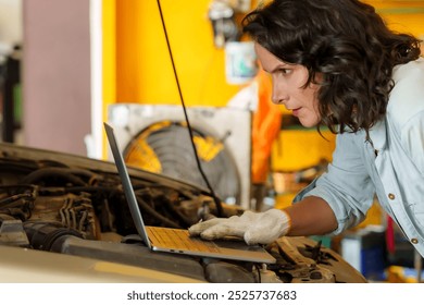 Focused Caucasian female mechanic working on a laptop, placed on a car engine in an automotive workshop, wearing light blue shirt and white gloves, displaying modern technology use in car diagnostics. - Powered by Shutterstock