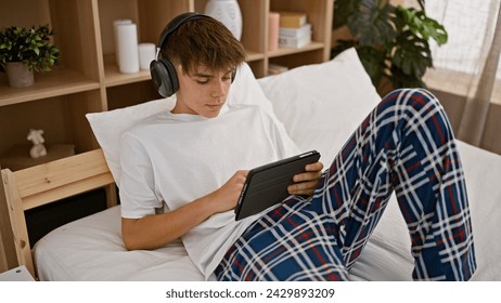 A focused caucasian teenage boy in pajamas engaging with a tablet in his bedroom while wearing headphones - Powered by Shutterstock