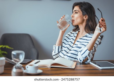 A focused Caucasian woman pauses to drink water in her modern workspace adorned with the latest gadgets and a time-keeping hourglass. - Powered by Shutterstock