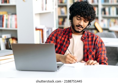 Focused Clever Arabian Or Indian Male Student, With Headset, Sits At Table With Laptop In University Library, Preparing For Exam, Studying Information, Listens Lecture, Concentrated Takes Notes