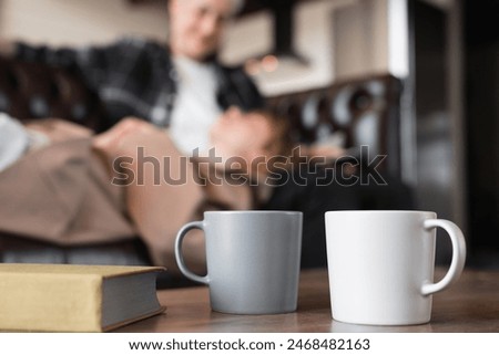 Focused close up image of two tea mugs cups with a book on coffee table and two young lovers in background. Spending time together. Love and passion concept. Pastime with spouse girlfriend boyfriend