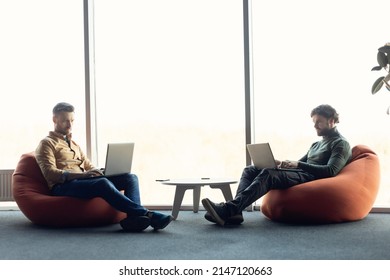 Focused Colleagues Sitting In Bean Bag Chairs, Working With Laptop Computers At Open Space Office, Copy Space. Two Serious Professionals Using Pc Devices, Coworking Together At Modern Company