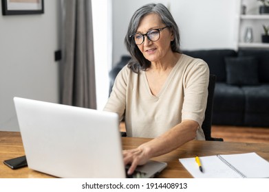 Focused and confident senior woman in glasses using a laptop for remote work sitting at the desk at home. Concentrated aged woman typing mails answers, messaging online