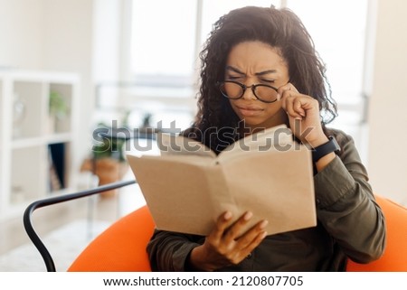 Focused confused young black lady trying to read paper book, squinting to see more clearly, wearing glasses, having difficulties seeing text because of vision problems, sitting on pouf in living room