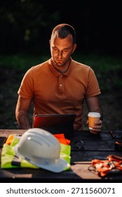 Focused construction engineer using tablet computer and drinking coffee at table with safety equipment outdoors