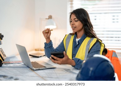 A focused construction professional woman reviewing project plans with a laptop and phone at a bright work desk - Powered by Shutterstock