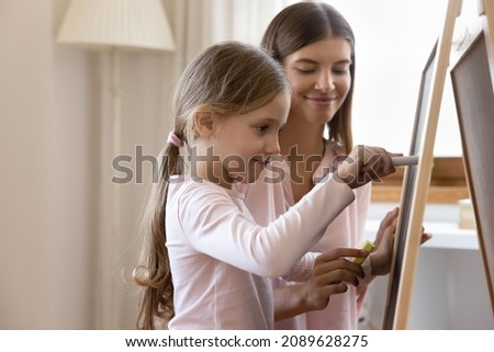 Focused cute girl kid learning to write, drawing with chalk on chalkboard under control of daycare teacher. Happy mom and daughter kid studying in game at home, enjoying education activity