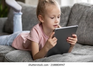 Focused Cute Preschooler Girl Watching Movie, Animated Films, Cartoons On Tablet Computer. Child Girl Lying On Couch In Living Room, Using Digital Pad For Video Call, Holding Device Close To Face