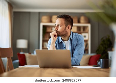 A focused, deep in thought male freelance copywriter looking aside with his hand on his chin sitting in front of a laptop having a writer's block.