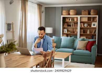 A focused, deep in thought male freelancer with his hand on his chin, looking at a project on a paper sheet while sitting at a table in front of a laptop in a home-based office. - Powered by Shutterstock