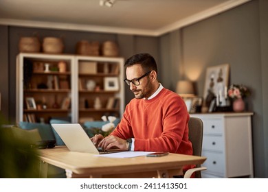 A focused and diligent adult male online content writer typing on his laptop and sitting at a wooden desk in his home office.