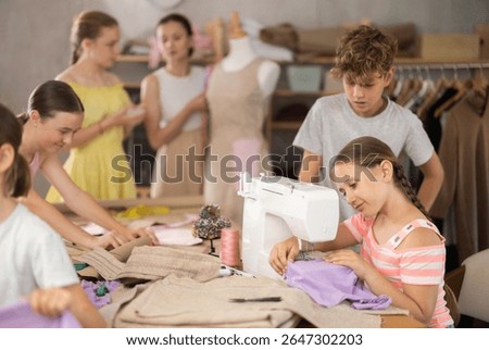 Focused and diligent ten year old girl carefully sewing on machine during hands-on class, while classmates preparing patterns and fabric under guidance of female teacher..