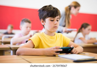 Focused Diligent Tween Schoolboy Sitting At Lesson In Classroom, Using Mobile Phone