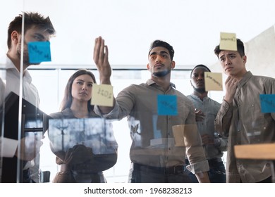 Focused Diverse Group Of Business People Discussing Idea Or Task, Looking At Post It Sticky Notes On Glass Wall. Serious Team Developing Work Plan In Creative Corporate Office At Stand Up Meeting