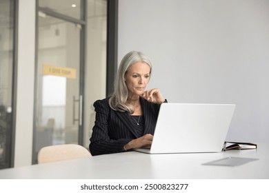 Focused elder company executive woman in suit working at laptop in meeting room alone, sitting at large white office table, typing, using professional application. Business leader candid portrait - Powered by Shutterstock