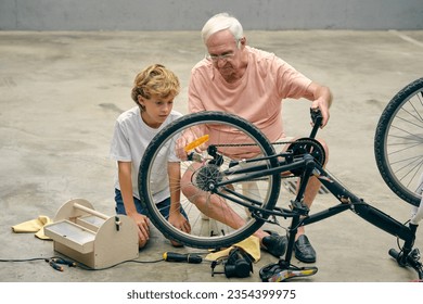 Focused elder man with grandson repairing bicycle wheel while sitting on concrete floor - Powered by Shutterstock