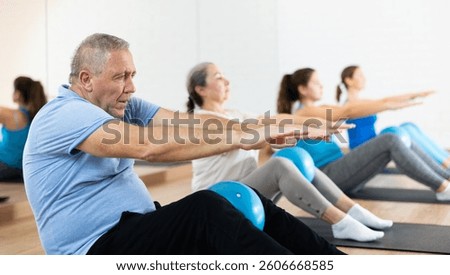Focused elderly man doing sit-up with bender ball to strengthen abs muscles during group pilates class in fitness studio