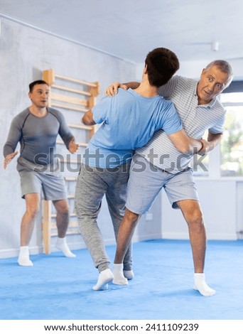 Focused elderly man practicing close combat techniques in sparring with young opponent in self-defence training room under guidance of experienced instructor..