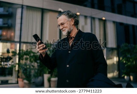Focused elderly professional texting on smartphone, side view of mature man checking financial news or trading stocks near glass office center, copy space, candidate photography, digital connectivity.