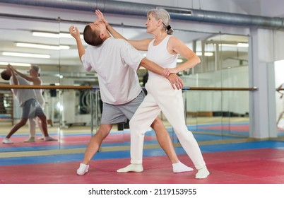Focused elderly woman practicing basic self-defense techniques while training in gym with male partner, performing palm heel strike in chin