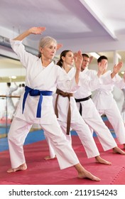 Focused elderly woman wearing kimono standing in attacking stance, practicing movements of close combat punches in training room during group martial arts workout..