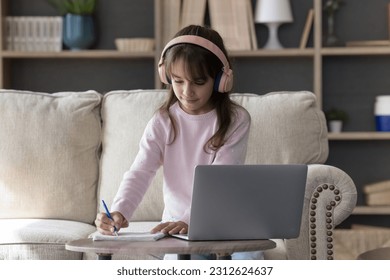 Focused elementary school student kid girl in pink headphones watching teachers learning webinar on laptop, writing notes, lesson, tutorial summary in copy book, studying on Internet from home - Powered by Shutterstock