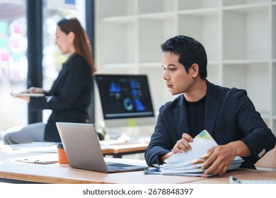 Focused employee analyzing documents and using laptop, with colleague working in background in bright office space