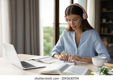 Focused engaged student girl in big headphones studying foreign language, listening audio lesson at laptop, reading notes out loud, doing exercises from open book, school, college homework task