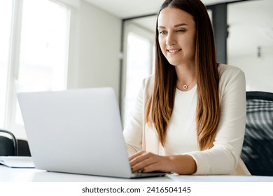 Focused and engaged a young businesswoman types away on a laptop at an office desk, focused female office employee responding emails sitting on the comfy well-organized workplace - Powered by Shutterstock