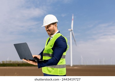 A focused engineer examines his laptop with a wind turbine in the background under a clear sky. - Powered by Shutterstock