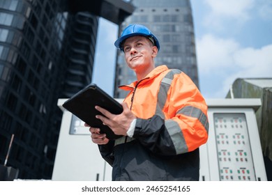 Focused engineer in high-visibility jacket and blue hard hat using a tablet for operational management at an industrial site with control panels.