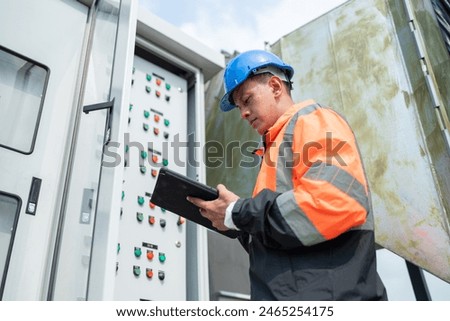 Focused engineer in high-visibility jacket and blue hard hat using a tablet for operational management at an industrial site with control panels.