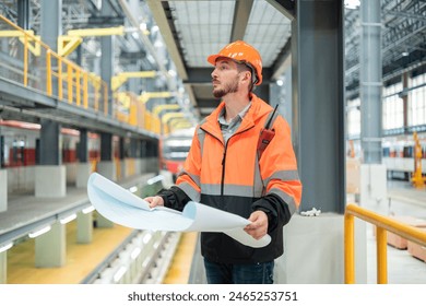 Focused engineer in reflective safety jacket examining blueprints in a busy train maintenance workshop. - Powered by Shutterstock