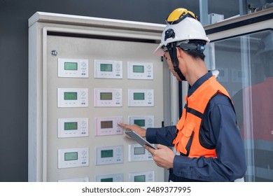 A focused engineer in safety gear examines an electrical control panel while holding a digital tablet. - Powered by Shutterstock