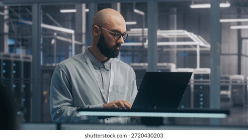 Focused IT Engineer Using Laptop in Server Hall. Man in Light Shirt Standing and Working Intently, Background Shows Server Racks Modern Data Center. Concept On Site Diagnosis and System Maintenance.