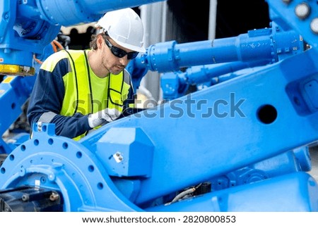 A focused engineer wearing a white hard hat and yellow safety vest uses a tablet to inspect a large blue robotic arm in an industrial workshop.