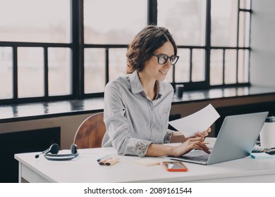Focused European businesswoman sitting at home office holds financial documents looks on laptop screen while typing some data. Serious young lady works remotely online for financial company management