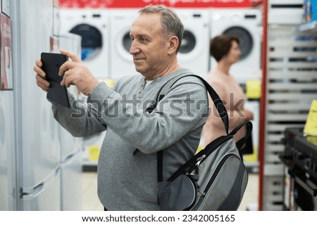 Focused European man who came to an electronics and household appliances store for a purchase takes pictures with a mobile ..phone camera of a sign with a price tag on the refrigerator