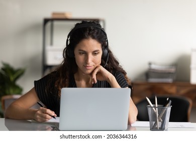 Focused female adult student in headphones using laptop, attending online lesson, virtual class, listening webinar, watching training, writing notes, making video call. Remote learning on internet - Shutterstock ID 2036344316