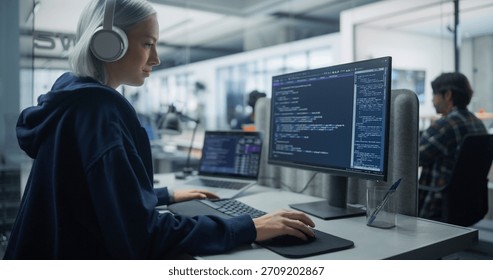 Focused Female Coder With Headphones Programming on Dual Monitors in a Modern Office. Engaged in Software Development, Optimizing Digital Solutions With Advanced Technology for Efficient Innovation.