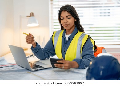 Focused female construction manager reviewing plans and using smartphone in modern office space during daytime - Powered by Shutterstock